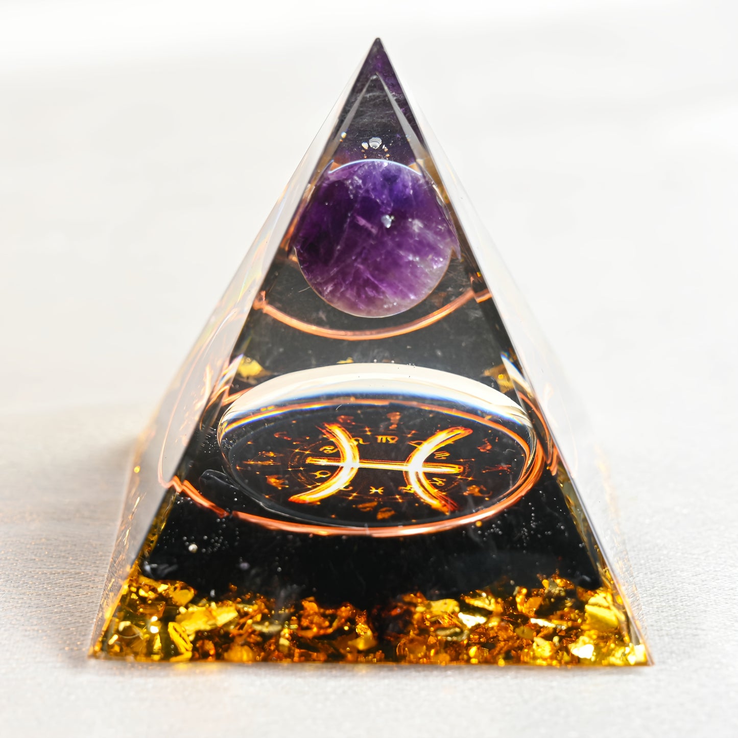 An orgone pyramid featuring a Pisces zodiac symbol, amethyst sphere, copper coil, black crystals, and golden foil chips.