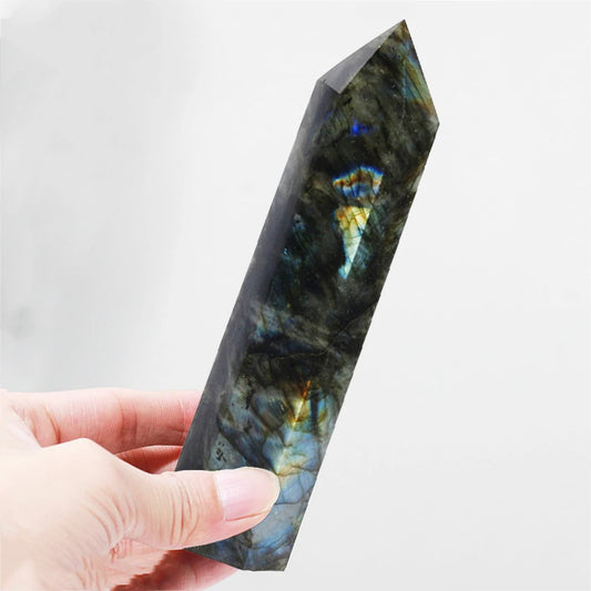Close-up of a hand holding a polished labradorite crystal tower showcasing iridescent blue and gold flashes against a light background.