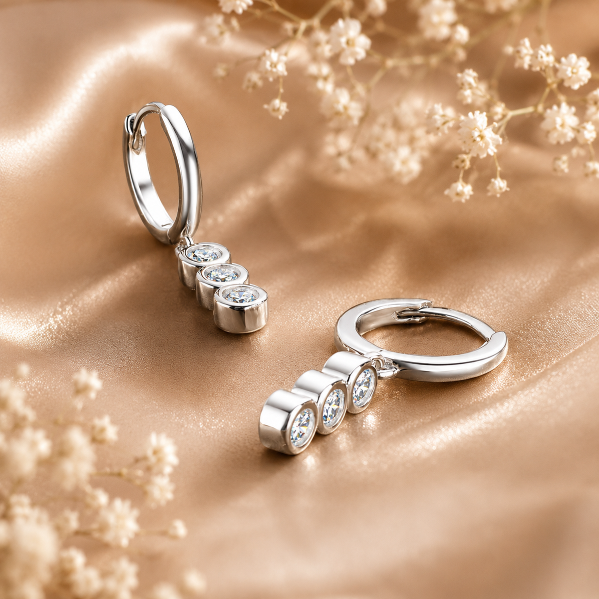 Silver triple-drop moissanite hoop earrings displayed on champagne satin with delicate flowers