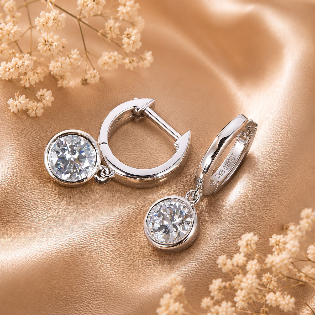 Silver bezel drop moissanite hoop earrings displayed on champagne satin with dried flowers