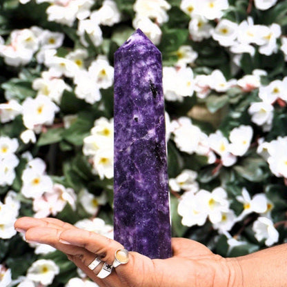 Hand holding a large Lepidolite tower against a background of white flowers, highlighting the stone’s rich violet colour and natural texture for anxiety relief and emotional balance.