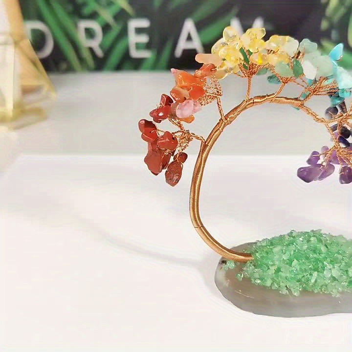 Rainbow chakra crystal bonsai tree with natural stone base, displayed on white table with blurred inspirational text in background.