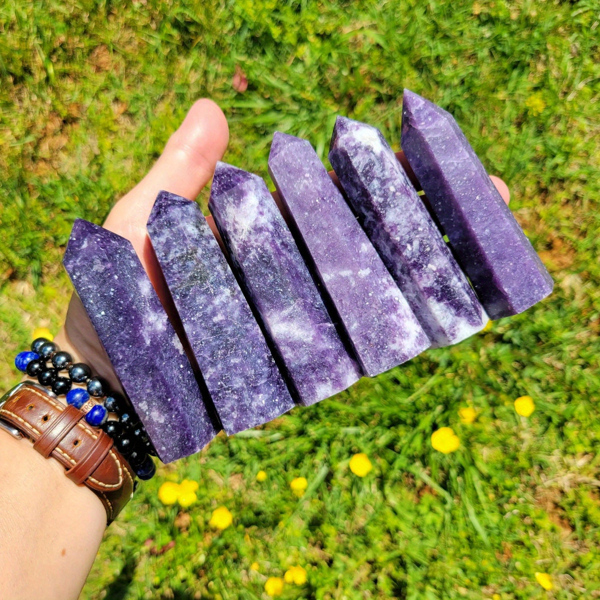 Six Lepidolite tower crystals displayed in hand outdoors, surrounded by green grass and yellow flowers, promoting peace, balance, and chakra alignment.