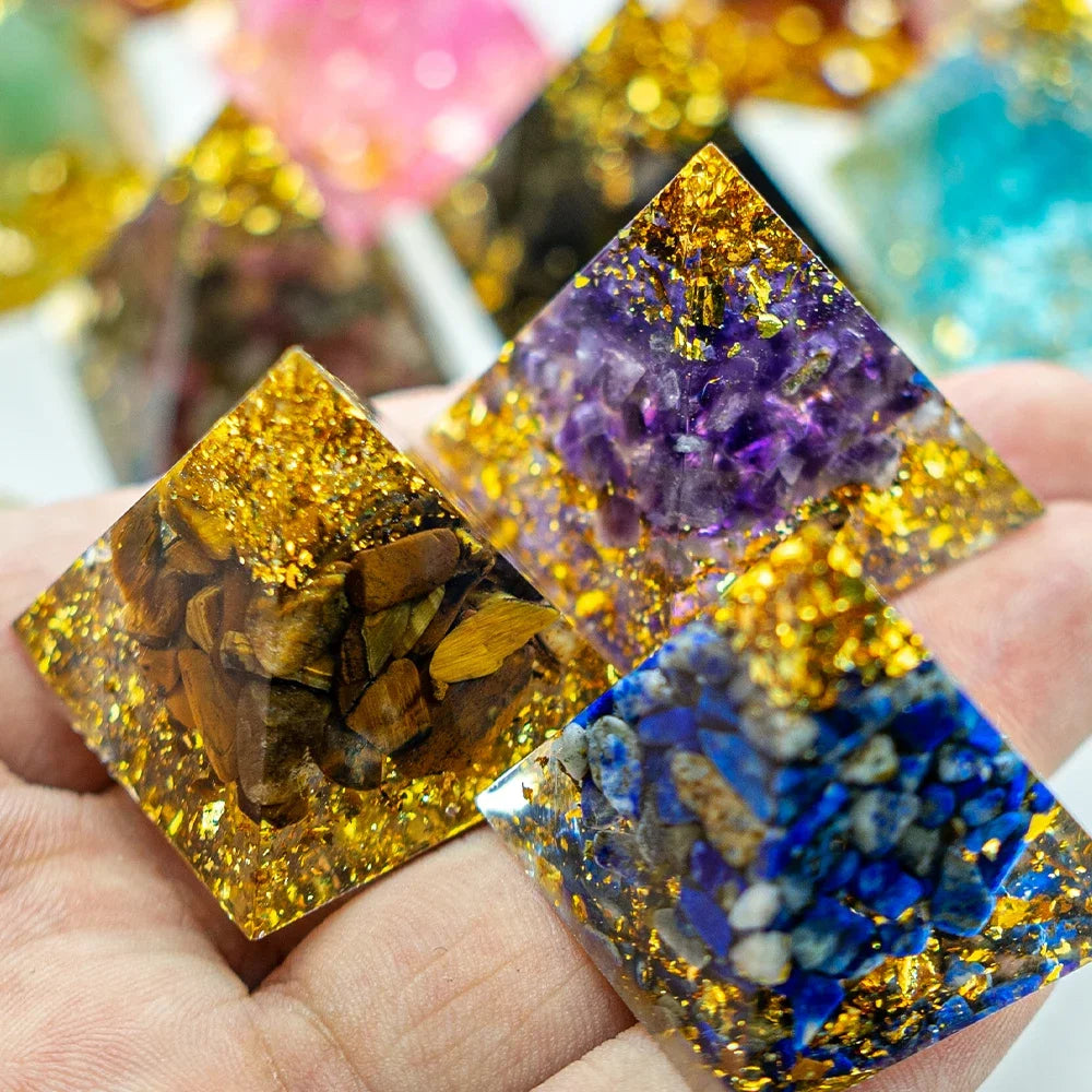 Close-up of a hand holding three mini 3cm orgone pyramids, each featuring different crystal types—tiger’s eye, amethyst, and lapis lazuli—encased in clear resin with shimmering gold foil, designed for energy healing and EMF protection.