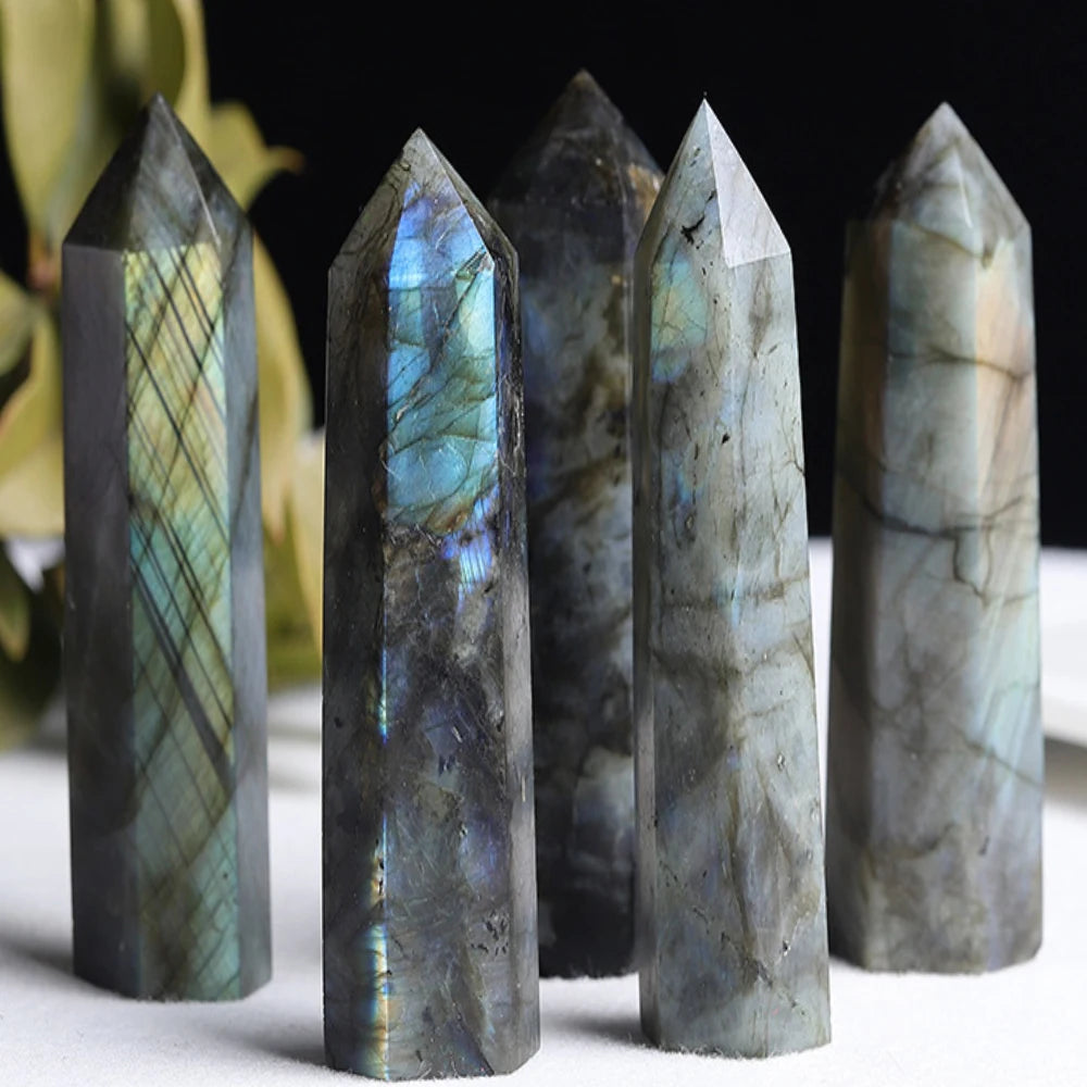 Five labradorite crystal towers arranged upright on a light surface with leaves in the background, highlighting their natural shimmer and colour variation.