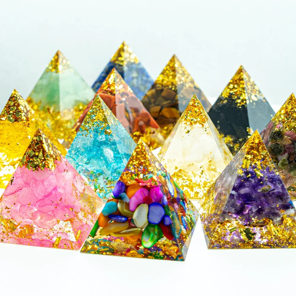 A vibrant display of twelve handcrafted 3cm orgone pyramids, each featuring unique crystal chips, colourful stones, gold foil, and clear resin, arranged in rows on a white background. Designed for chakra healing, energy balancing, and EMF protection.