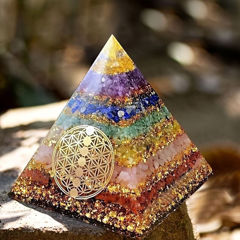 Handcrafted 7 chakra orgone pyramid featuring layered crystals, gold flakes, and a golden Flower of Life symbol, placed on a textured stone surface with warm natural sunlight and a blurred earthy background.